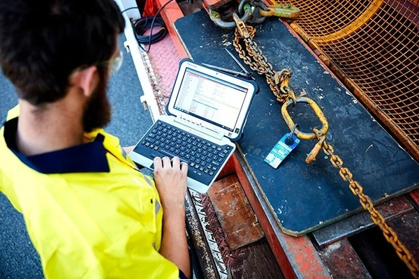 A bearded man wearing a high visibility vest uses a computer with open inspection software. He is standing before a workbench with a lifting chain connected by a master link.